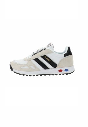 Baskets beiges et blanches avec des rayures noires, dessus en suède et en mesh, semelle texturée et accents colorés sur le talon en rouge et bleu.