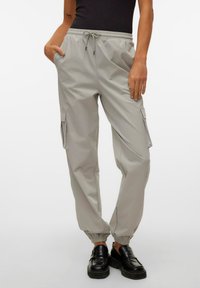 Pantalons cargo gris clair avec des poignets et une taille élastiques. Comprend des poches latérales et une texture lisse. Associés à des chaussures noires.