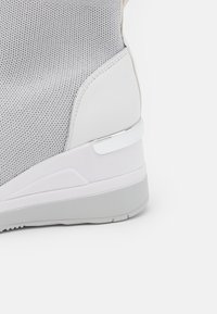 Šedá sportovní bota s síťovaným svrškem a bílými syntetickými akcenty. Má texturovanou podrážku a kovový detail na patě.