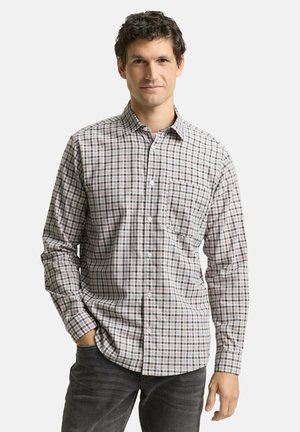 Mann trägt ein langärmliges kariertes Hemd mit Knöpfen und dunkle Jeans, steht mit einer Hand in der Tasche vor einem schlichten Hintergrund.