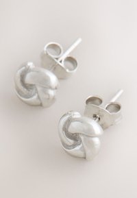Pair of small silver stud earrings shaped like intertwined knots on a light background.