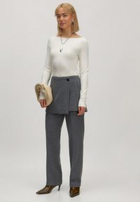 Grijze tailored broek met een asymmetrische voorkant, gecombineerd met een lange mouwen witte top, en een beige accessoire vasthoudend. Bruine puntige hakken.