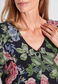 Blumenbluse mit V-Ausschnitt, dunkler Hintergrund. Ausgestattet mit rosa und blauen Blumen, grünen Blättern und schwarzen Knöpfen an der Vorderseite. Glatte Stofftextur.