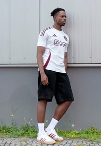Hvid atletisk skjorte med maroon accenter, sorte shorts og hvide sko med røde striber. Står mod en grå væg med grønne planter nedenfor.