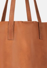 Tote-Bag aus tanfarbenem Leder mit zwei flachen Griffen, sichtbarer Naht und eingeprägtem Logo. Glatte Textur mit subtilen Maserungen.