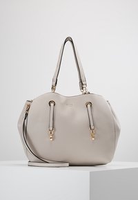 Vaalean beige nahkainen käsilaukku kultasävyisillä yksityiskohdilla, joissa on kaksi yläkahvaa ja irrotettava olkahihna, lepäilee valkoisella jalustalla.