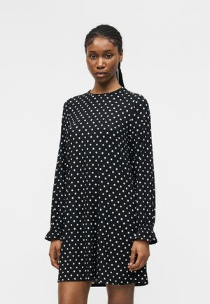 Kvinde med fletninger iført en sort langærmet kjole med hvide prikker, stående mod en ensartet lys baggrund.