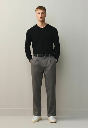 Ung mand står mod en ensfarvet væg iført en sort sweater, grå bukser, hvide sneakers, med hænderne i lommerne.