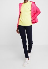 Jaskrawo różowa, ocieplana kurtka na żółtym swetrze z dzianiny, zestawiona z ciemnymi dżinsami i białymi trampkami. Posiada ściągacze przy mankietach oraz detal z logo.