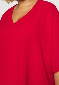 Haut rouge à col en V en tissu lisse, avec des manches courtes et une coupe décontractée, mettant en valeur un design simple et sans décorations.