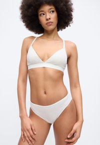 Νεαρή γυναίκα με κατσαρά μαλλιά που φοράει λευκό μινιμαλιστικό σουτιέν και εσώρουχα, στέκεται μπροστά σε ένα απλό φωτεινό φόντο.