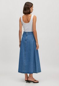 Denim maxi rok in medium blauw, met een hoge taille, A-lijn silhouet en subtiele stikdetails. Gecombineerd met een witte tanktop.
