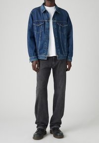 Muž v modré džínsové bundě přes bílou košili, tmavě šedých džínách a černých šněrovacích botách, stojící proti jednoduchému světlému pozadí.