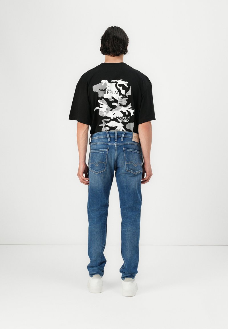 Camiseta oversize negra con un patrón de camuflaje blanco en la parte posterior, combinada con jeans de mezclilla azules y zapatillas blancas.