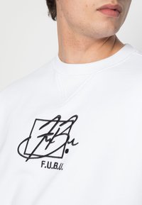 Witte sweatshirt met ronde hals en geborduurd zwart logo, met gestileerde tekst en een doosontwerp op de borst. Zachte stof, textuurafwerking.