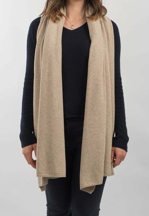 Vrouw draagt een lange beige sjaal over een zwarte trui en zwarte broek, met een ketting met een hartvormige hanger, staand tegen een effen achtergrond.