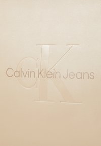Bežasti teksturirani površina s uzdignutom oznakom "CK Calvi Klein Jeans" u minimalističkom fontu s glatkom završnom obradom.