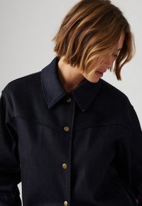 Marine denimjakke med messingknapper, spiss krage og brystsømdetaljer; materialet ser robust og teksturert ut.