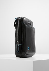 Borsa in pelle nera con texture liscia e cuciture blu a contrasto, dotata di chiusura zip e tasca frontale. Forma rettangolare con angoli arrotondati.