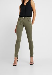 Olive green skinny jeans with front zip pockets, smooth fabric, and a fitted design paired with a black sleeveless top.