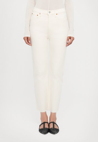 White, high-waisted jeans with a straight fit, frayed hem, and copper-colored hardware. Paired with black, ribbed flats.