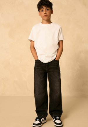 Niño de pie con las manos en los bolsillos, vistiendo una camiseta blanca, pantalones vaqueros negros holgados y zapatillas en blanco y negro, frente a un fondo beige.