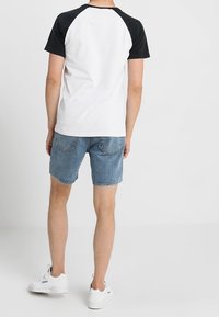 Camiseta raglán en blanco y negro de manga corta, combinada con pantalones cortos de mezclilla azul claro. Zapatillas blancas completan el atuendo, visto desde atrás.