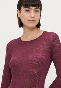 Hosszú ujjú felső sötét burgundy színben, áttetsző anyagból készült, finom tigriscsíkos mintával, kerek nyakkivágással és testhez simuló fazonnal.