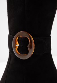 Botín de ante negro con una amplia banda y una hebilla circular de color ámbar, además de detalles en plata, que destaca por su suave textura y diseño elegante.