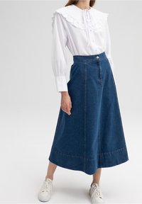 Bílá blůza s volánkovým límcem a zapínáním na knoflíky v kombinaci se sukní z modrého džínu střední délky, která má přední panely a jeden knoflík.