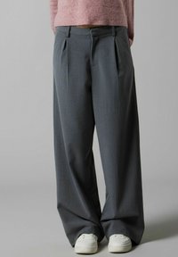 Pantalons larges en tissu gris foncé avec des plis ; associés à des baskets blanches. Texture lisse, design ajusté et coupe décontractée.