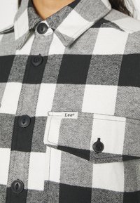 Black and white checked flannel shirt with pointed collar, black buttons, and a chest pocket featuring a Lee® label. Soft texture.