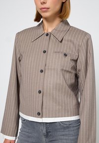 Bruin gestreept cropped jack met lange mouwen, voorzien van een puntige kraag, zwarte knopen en een voorke pocket met een subtiele accent.