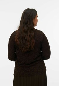 Rjava čipkasta bluza z dolgimi rokavi, z nazobčanim robom. Tkanina ima teksturiran cvetlični vzorec, zadnji pogled pa prikazuje sproščene valove v laseh.