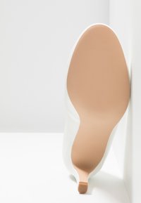 Chaussure à talons hauts blanche montrée par la semelle avec un dessous beige lisse et un petit talon sur fond blanc.