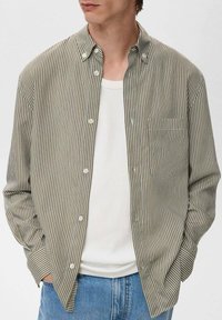 Camisa de botones a rayas en tonos apagados de verde y marrón, con un bolsillo en el pecho y un corte relajado. Lleva una camisa blanca debajo.