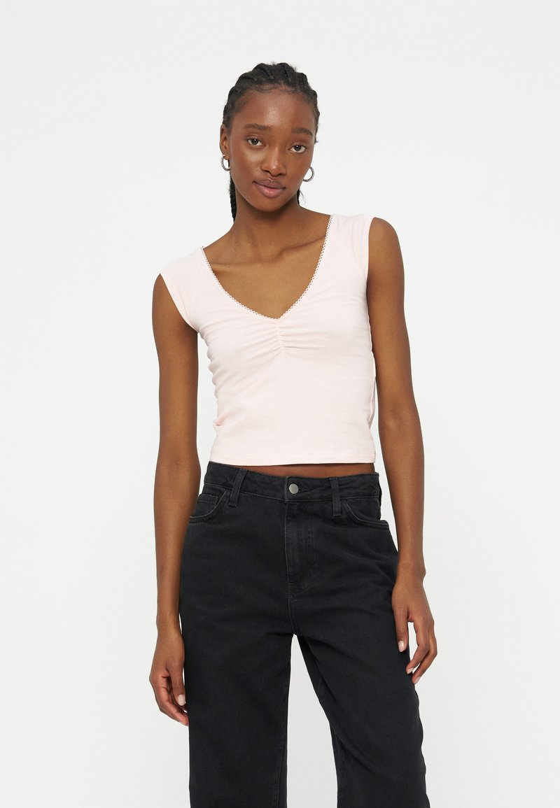 Νεαρή γυναίκα με πλεξούδες, φοράει ανοιχτό ροζ αμάνικη μπλούζα με πιέτες και μαύρο παντελόνι ψηλόμεσο, στέκεται μπροστά σε λευκό φόντο.