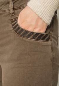 Jeans de mezclilla marrón con un bolsillo texturizado y acentos de rayas oscuras. Costuras visibles y un acabado de tela natural.