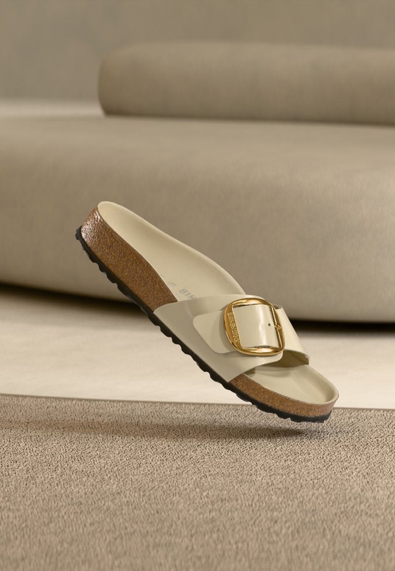 Μπεζ σανδάλι Birkenstock με χρυσή αγκράφα και φελό πάτο, ακουμπά σε υφασμένο μπεζ χαλί με φόντο μπεζ καναπέ.