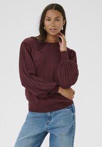 Pull en tricot bourgogne avec des manches longues bouffantes et côtelées, col rond, associé à un jean bleu clair. Texture lisse et coupe décontractée.