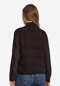 Kvinna sedd bakifrån iklädd en svart cable-knit ärmlös väst över en svart långärmad tröja och blå denimjeans.