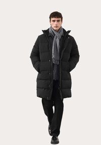 Schwarze Daunenjacke mit gestepptem Design, Frontreißverschluss und Druckknöpfen. Kombiniert mit einem grauen Schal, über einem dunklen Oberteil und schwarzen Hosen.