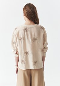 Blusa de textura cremosa con sutiles rayas verticales y patrones florales bordados en marrón, combinada con pantalones beiges holgados.