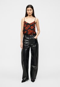 Μαύρο crop top με κόκκινο μοτίβο τριαντάφυλλου, συνδυασμένο με μαύρο, γυαλιστερό, ψεύτικο δερμάτινο παντελόνι με φαρδιές παντελόνια και ψηλή μέση. Το μοντέλο στέκεται με τα χέρια στις τσέπες.