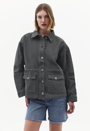 Junge Frau trägt eine dunkelgraue, übergroße Jacke mit Knöpfen und Klapptaschen sowie hellblaue Jeans-Shorts und steht vor einem schlichten weißen Hintergrund.