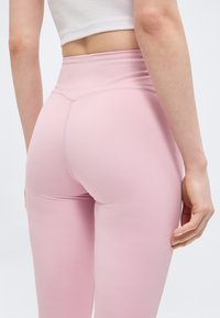 Γυναίκα που φοράει ψηλόμεσα ανοιχτό ροζ κολάν και λευκό κοντό τοπ, απεικονιζόμενη από πίσω μπροστά σε απλό ανοιχτό φόντο.