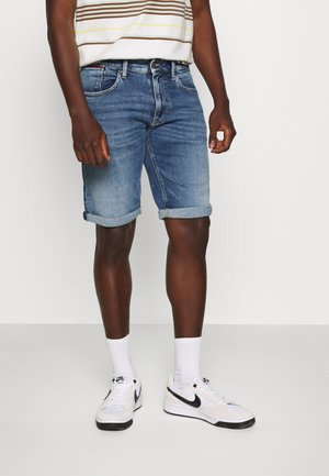 Mand iført opkrogede blå denimshorts, hvide ankelstrømper, hvide Nike-sneakers med sort swoosh og en hvid stribet skjorte, stående på gråt gulv.
