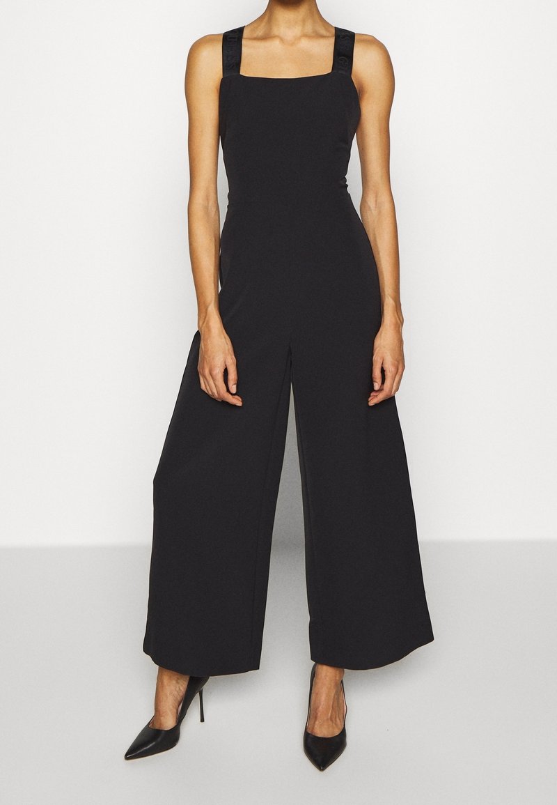 Combinaison noire à jambes larges, encolure carrée et bretelles en satin. Tissu lisse avec une coupe ajustée. Portée avec des talons noirs pointus.