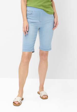 Kvinde iført lyseblå knælange denimshorts, grøn top og hvide åbne tå-sandaler, stående mod en hvid baggrund.