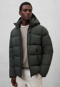 Mørkegrøn dunjakke med hætte, med en lynlåslomme på brystet, quiltet design og tekstureret stof. Båret over en lyse trøje.